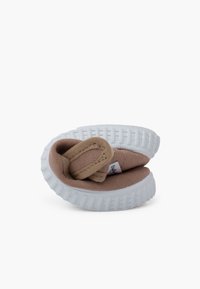 Chaussure en toile marron avec une semelle en caoutchouc blanc, dotée d'une sangle boucle et d'un intérieur doux. Forme arrondie, design minimaliste, sans motifs visibles.