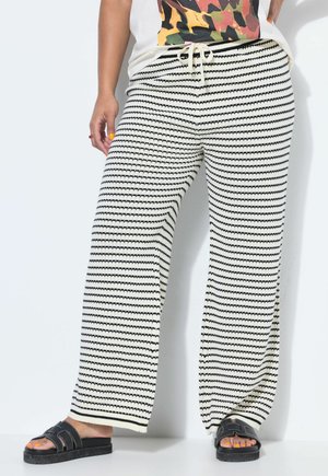 Osoba nosząca szerokie spodnie w białe i czarne paski z sznurkiem w talii, czarne klapki i kolorową, wzorzystą bluzkę.