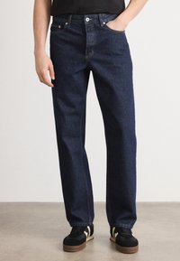 Temno modri denim hlače s ravnim rezom, načrtom s petimi žepi in vidnimi šivi. Obute s črnimi športnimi copati z kremnimi detajli.