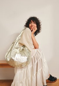 Velika, metalno srebrna hobo torba z mehko teksturo, ki ima enotno naramnico in sproščen dizajn. Oblečena z lahkotno, večplastno krilo.