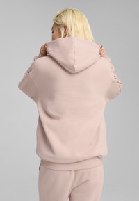 Lyserød hoodie med en rummelig hætte, afslappet pasform og ribbede manchetter. Materialet ser blødt ud med minimal syning og ingen synlige mønstre.