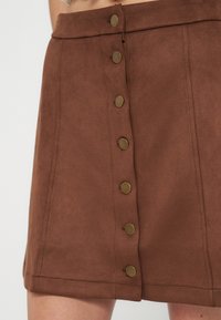 Jupe en daim marron avec une rangée verticale avant de six boutons-pression en laiton, portée à la taille, montrant la partie supérieure de la cuisse.