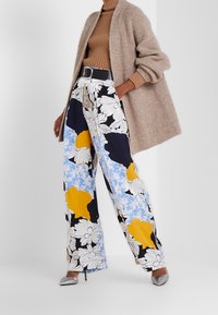 Wijde bloemenbroek in blauw, geel en zwarte patronen; gecombineerd met een geribde bruine top en een beige gebreide cardigan. Zilveren hak schoenen.