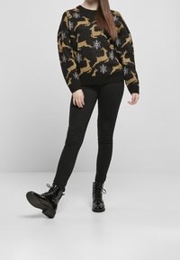 Frau trägt einen schwarzen Pullover mit goldenem Rentier und weißen Schneeflocken, schwarze enge Hose und glänzende schwarze Schnürstiefel.