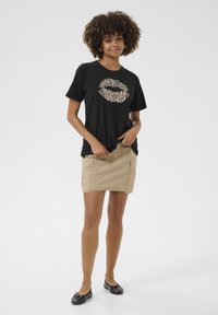 Zwarte T-shirt met een lippenafbeelding in leopardprint, gecombineerd met een beige cargorok. Model draagt zwarte ballerina's. Eenvoudig ontwerp, ontspannen pasvorm.