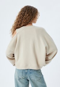 Felpa oversize beige con scollo ampio e dettagli sulle spalle scoperte, realizzata in tessuto morbido, abbinata a jeans in denim azzurro chiaro.