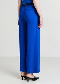 Pantalon à jambes larges en tissu bleu vif, présentant un design épuré avec une ceinture élastique et un détail poche arrière. Des mules à talons noires sont portées.