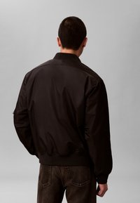 Man draagt een zwarte bomberjack en donkere spijkerbroek, kijkt weg en staat tegen een effen lichtgrijze achtergrond.