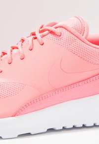 Gros plan d'une chaussure de sport rose avec tige en mesh et synthétique, semelle texturée et lacets, reposant sur une surface blanche.
