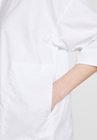 Camicia di cotone bianca con una vestibilità rilassata, dotata di una grande tasca laterale e maniche arrotolate. Tessuto liscio e design minimale.