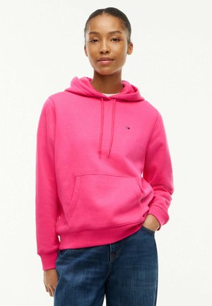 Femme portant un sweat à capuche rose vif avec poche avant et un jean bleu, debout avec une main dans la poche sur un fond uni.