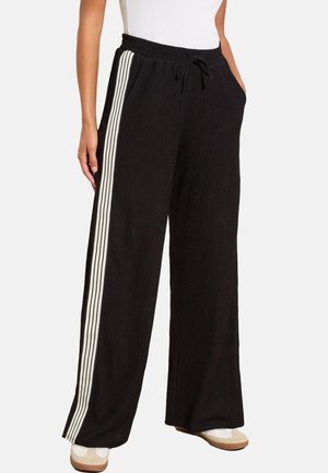 Kvinde iført løstsiddende sorte bukser med hvide lodrette striber på siderne, elastisk talje med snøre, hvid top og beige sko.