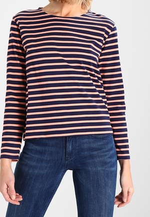 Femme portant un haut à manches longues à rayures horizontales bleu marine et rose, associé à un jean en denim bleu, debout devant un fond uni.