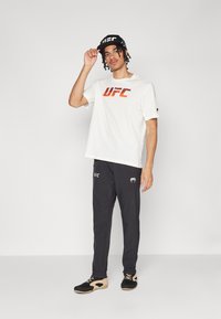 Balts T-krekls ar oranžu "UFC" logo, melni sporta bikses ar logo, melna cepure ar baltu logo un melnas kurpes ar brūnām akcentiem.