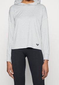 Vaaleanharmaa hupullinen collegepaita, jossa on pitkät hihat, löysä muoto ja pieni musta logo helman kohdalla. Yhdistetty tummiin ribattuihin trikoot. 