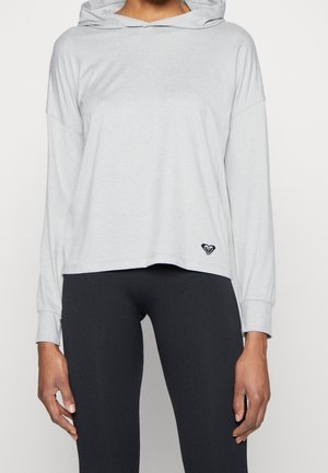 Jasnoszara bluza z kapturem, z długimi rękawami, luźnym krojem i małym czarnym logo na dolnym brzegu. Połączona z ciemnymi, prążkowanymi legginsami.