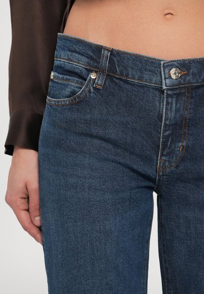 Közelkép egy személyről, aki középmagas derekú kék farmernadrágot visel narancssárga varrással és ezüst gombbal, sötét színű hosszú ujjú felsővel párosítva.