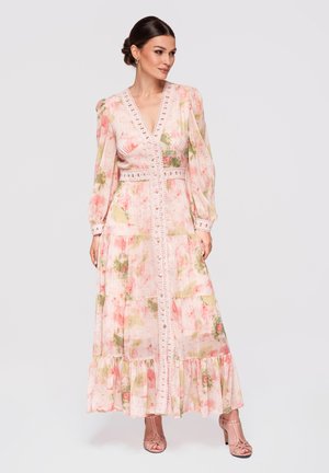 Vrouw draagt een lange, bloemrijke, pastelroze jurk met kanten afwerking, opgeblazen mouwen en een geplooide zoom met laagjes, gecombineerd met open-toe hakken, staand tegen een effen achtergrond.