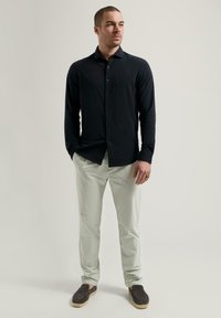 Svart langermet skjorte med knapper, parret med lysegrønne bukser og mørke slip-ons med jutesåle. Enkelt, tilpasset design.