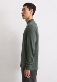 Junger Mann in grünem Langarmshirt und dunkler Hose, der seitlich gegen eine schlichte helle Wand steht.