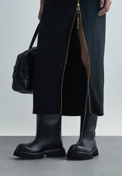 Personne portant des bottes en cuir noir jusqu'au genou avec des fermetures éclair centrales, une jupe noire avec une fente à fermeture éclair dorée, tenant un sac noir.