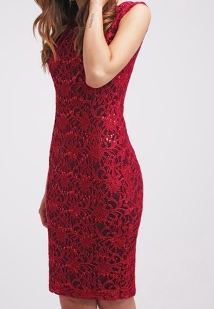 Woman wearing a sleeveless, knee-length red lace dress with floral patterns and sequins against a plain light background.