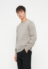 Light grey textured sweater with a crew neck and ribbed cuffs, worn over black trousers. Features a relaxed fit and soft fabric.