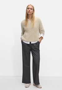 Beżowy, puszysty sweter z dekoltem w serek, biała koszula pod spód, szare spodnie w prążki o szerokim kroju, oraz beżowe buty. Modelka stoi z rękami w kieszeniach.
