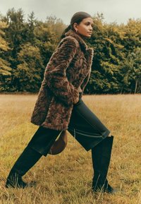 Mākslīgās ādas brūns mētelis, melna ādas svārki, zābaki līdz ceļgaliem melnā krāsā; modele tur brūnu rokassomu zāliena laukā ar meža ainavu fonā.