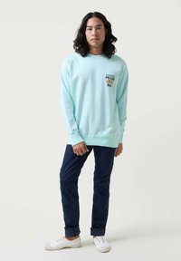 Sudadera de color verde claro con un pequeño logo en el pecho. Combinada con pantalones azul marino oscuros y zapatillas blancas tipo slip-on. Textura suave evidente.