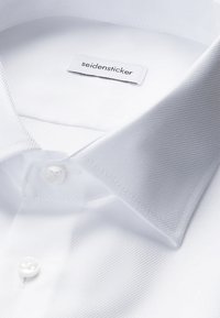 Camicia bianca con colletto aperto, bottoni bianchi visibili e etichetta del marchio "seidensticker" all'interno del colletto.