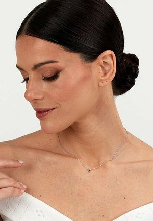 Silberkette mit einem kleinen, detaillierten Anhänger in Herzform, auf nackter Haut getragen, hebt den Halsbereich elegant hervor.
