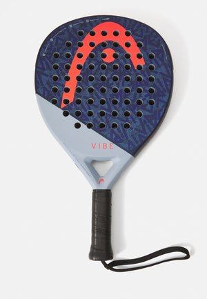 Pala de pádel con una superficie texturizada azul y verde, acentos rojos, perforaciones circulares y un mango de goma negro con correa para la muñeca.