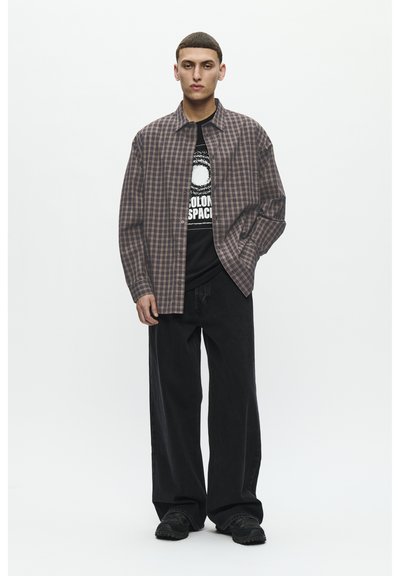 Cămașă cu print tartan în nuanțe de brun și negru, purtată peste un tricou grafic negru, asortată cu pantaloni negri lejeri și încălțăminte închise la culoare. Design simplu.