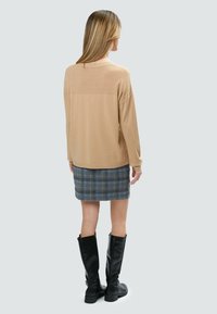 Beige gestrickter Pullover mit strukturiertem Muster auf den Schultern, kombiniert mit einem grauen karierten Rock. Schwarze kniehohe Stiefel vervollständigen das Outfit.