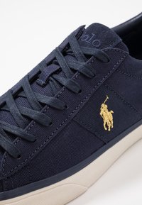 Granatowe trampki z płótna z wyszywanym złotym logo, płaskimi sznurówkami i teksturowanym bocznym panelem. Mają kremową gumową podeszwę.