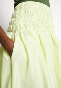 Jupe jaune clair avec une taille smockée, dotée de poches latérales et d'un design volumineux et texturé. Tissu lisse et légèrement brillant.