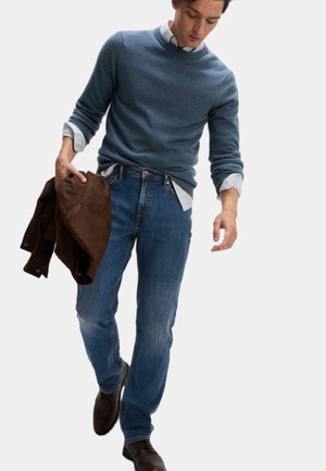 Mand iført blå sweater og jeans, holder en brun jakke i venstre hånd, går med hovedet hældt nedad mod en ensfarvet baggrund.