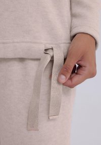 Beżowy sweter z teksturowym wykończeniem i ściągaczem. Szczyt ściagacza ma wzór w rybie łuski i metalowe akcenty na końcach.