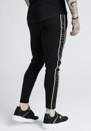 Mand iført sorte slim-fit bukser med skinnende sorte sidelommer og hvide striber, kombineret med sorte sneakers med hvide såler.