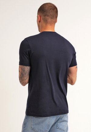 Mężczyzna z krótkimi włosami i wytatuowanymi ramionami, ubrany w prosty granatowy t-shirt i jasnoniebieskie jeansy, stoi tyłem do kamery na jednolitym tle.