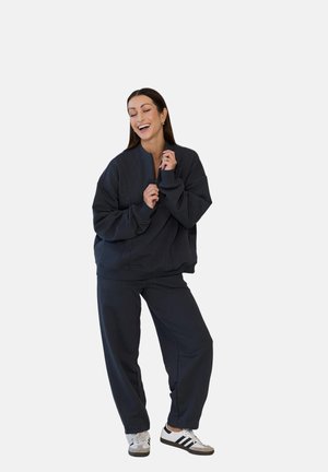 Kobieta uśmiechająca się z zamkniętymi oczami, ubrana w ciemną, za dużą bluzę i spodnie, białe tenisówki, stojąca na tle jednolitego białego tła.