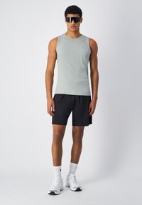 Helles grünes ärmelloses Sportshirt aus strukturiertem Stoff, kombiniert mit schwarzen Shorts. Modell trägt übergroße schwarze Sonnenbrille und weiße Sneakers.
