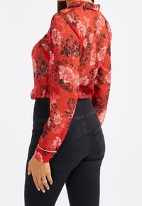 Blusa roja con flores y detalles negros, tela transparente, mangas largas y cuello abotonado, combinada con jeans ajustados negros.