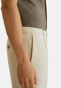 Beige linnen broeken met een getailleerde pasvorm, voorzien van voorplooien en een knoopzak aan de achterkant, gedragen met een grijze ribbel top met korte mouwen.