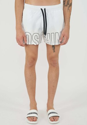 Homme portant un short de bain blanc avec un cordon noir et des lettres noires contourées sur le devant, debout pieds nus dans des claquettes blanches Emporio Armani.