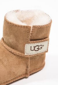 Botín de ante marrón hasta el tobillo con forro interior suave, detalles de costura y una etiqueta de logo UGG visible en el costado.