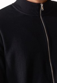 Giacca nera con zip, caratterizzata da un collo alto, un design elegante e una zip metallica. Texture morbida con una finitura in tessuto liscia.