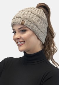 Beanie en tricot beige avec une texture côtelée et un motif diagonal, arborant un petit patch en cuir, porté avec un col roulé noir.
