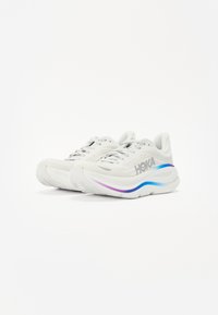 Baskets de course blanches avec une tige en maille, une semelle épaisse avec des accents dégradés de bleu et de violet, et le logo "HOKA" sur le côté.